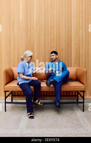 Junger männlicher Arzt, der mit einer älteren Kollegin spricht, während er auf dem Sofa in der Krankenhauslobby sitzt Stockfoto