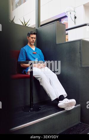 Volle Länge der reife männliche Gesundheitsarbeiter, die Pausenzeit mit Smartphone verbringen, während sie in der Krankenhauscafeteria sitzen Stockfoto