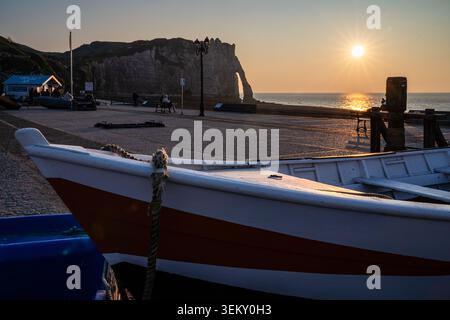 Blick von der Promenade Etretat mit dem Bug eines Holzbootes im Vordergrund. Im Hintergrund untergeht die Sonne über dem ruhigen Meer und beleuchtet die Stockfoto
