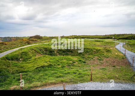 Weite Aussicht auf Pointe du hoc mit Kieswegen, die sich durch große grüne Grasbombenkrater schlängeln. Der Hintergrund zeigt Küstenbüsche, dist Stockfoto