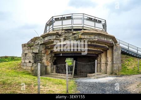 Vorderansicht eines Betonbunkers bei Pointe du hoc. Die Struktur verfügt über eine breite, offene Verteidigungsöffnung hinter einem Maschendrahtzaun, verwitterten Wänden und Stockfoto