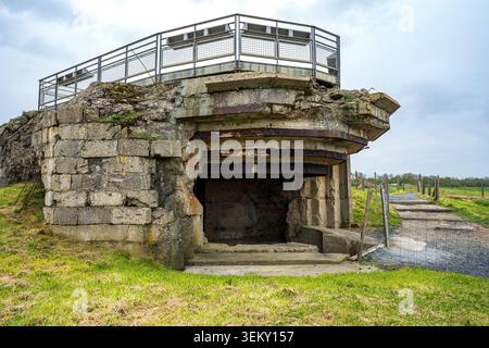 Vorderansicht eines Betonbunkers bei Pointe du hoc. Die Struktur verfügt über eine breite, offene Verteidigungsöffnung hinter einem Maschendrahtzaun, verwitterten Wänden und Stockfoto