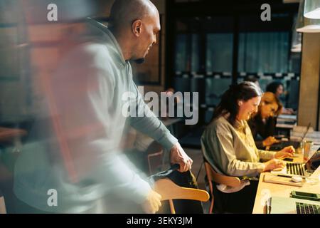 Geschäftsmann, der mit einer Kollegin spricht, die im Büro am Laptop sitzt und arbeitet Stockfoto