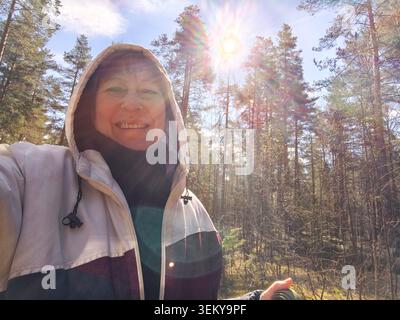 Eine Frau steht im Wald und lächelt, während sie ein Smartphone hält. Sonnenlicht zieht durch die Bäume, während sie ihre Zeit im Freien genießt. Stockfoto
