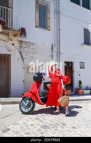 Junge Frau in trendigem Sommeroutfit posiert mit Roller im malerischen europäischen Dorf während der sonnigen Urlaubsreise. Stockfoto