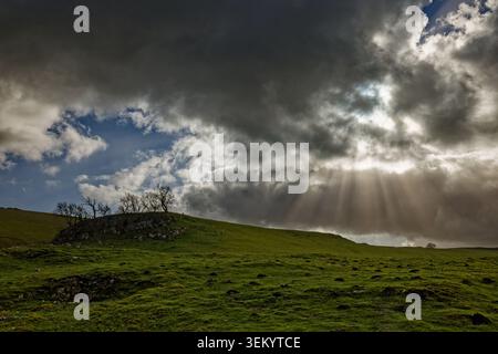 Die Sonnenstrahlen platzen durch dunkle Regenwolken in einer Hochgebirgslandschaft mit Bäumen auf einem felsigen Felsen, Castleton, Peak District, Derbyshire. Stockfoto