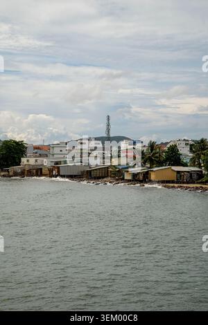Ein Blick vom Wasser einer kleinen vietnamesischen Küstenstadt mit bescheidenen Pfahlhäusern und Werkstätten entlang der felsigen Küste. Hinter ihnen stehen moderne W Stockfoto