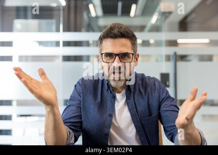 Frustrierter Mann, der mit offenen Handflächen die Arme zuckt und vor der Kamera spricht, Konflikte, Missverständnisse oder Unglauben in einer modernen Büroumgebung ausdrückt Stockfoto