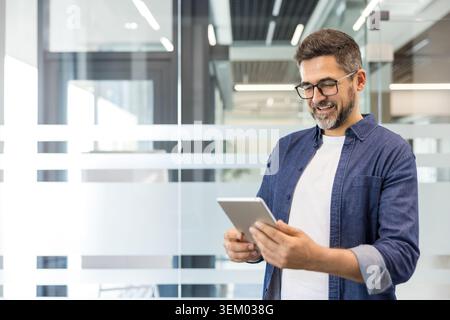 Ein lächelnder Mann mit Brille und Denim-Hemd steht in einem modernen Büro und ist mit einem digitalen Tablet beschäftigt, das Mobiltechnologie, Kommunikation und Geschäftsverbindungen repräsentiert Stockfoto