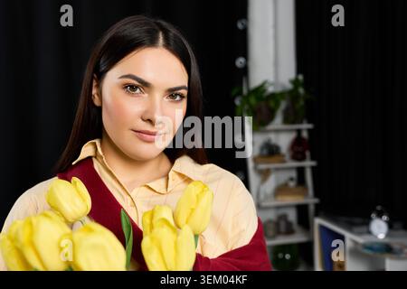Die charmante Frau lächelt zu Hause herzlich und wiegt leuchtend gelbe Tulpen in ihren Armen. Stockfoto