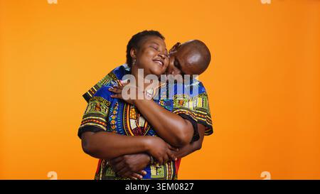 Süßes afroamerikanisches Paar, das sich im Studio umgibt, sich hält und ihre aufrichtigen Gefühle ausdrückt. Ethnisch-romantischer Mann umarmt seine Frau vor orangem Hintergrund. Stockfoto