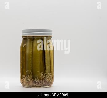 Ein Glas gefüllt mit ganzen Gurken, die in einer Mischung aus Salzlake und Gewürzen sitzen. Der Behälter ist mit einem Metalldeckel verschlossen. Die Einstellung ist einfach und hell und konzentriert sich auf die Gurken. Stockfoto