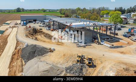 Auf einer Baustelle, auf der Metallträger für ein neues Gebäude installiert werden, sind die Arbeiter beschäftigt. Es sind schwere Maschinen sowie Materialstapel vorhanden. Das Gelände ist von offenem Land umgeben. Stockfoto