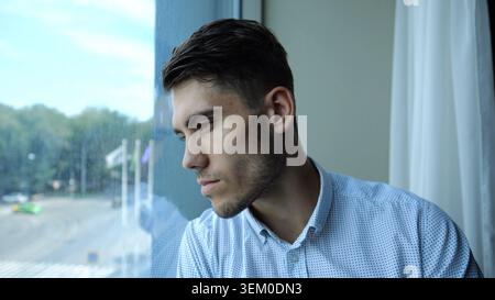 Porträt eines traurigen jungen Unternehmers, der in einem modernen Hotelzimmer oder Büro am Fenster sitzt. Unglücklicher Mann mit ernstem Ausdruck, der wegblickt Stockfoto