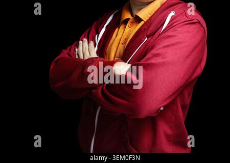 Der Mann steht mit überkreuzten Armen und trägt einen roten Hoodie. Ein hellorangefarbenes Hemd blickt von unten. Weiße Reißverschlusslinien betonen die Textur der Jacke. Hochwertige Fotos Stockfoto