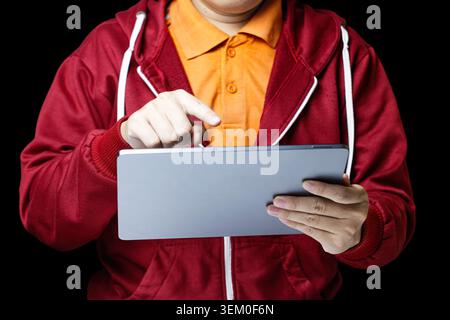 Person in rotem Hoodie mit einem digitalen Tablet. Tippen Sie mit dem Finger auf den Bildschirm mit fokussierter Absicht. Hellorangefarbenes Hemd steht im Kontrast zu dunklem Hintergrund. Ideal für Unternehmen und Bildungseinrichtungen. Hochwertige Fotos Stockfoto