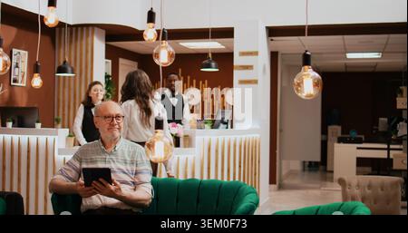 Ein älterer Mann überprüft die Verbindung des Tablets im Hotel und entspannt sich im Loungebereich, bevor er in den Ruhestand geht. Auf Online-Websites finden Sie Freizeiteinrichtungen und entspannende Aktivitäten. Stockfoto