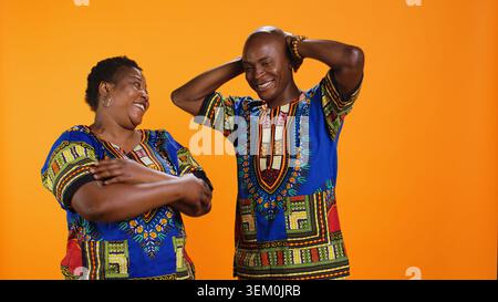 Fröhlicher Ehemann und Frau lachen zusammen vor der Kamera, haben Spaß mit albernen Witzen über orangem Hintergrund. afroamerikanisches Paar, das lustige Momente genießt, sorglose Leute lachen. Stockfoto