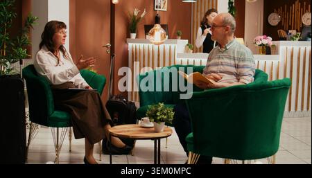 Ältere Ehepaare sprechen über Urlaubspläne im Lounge-Bereich des Hotels und warten darauf, das Zimmer einzuchecken. Ehemann und Ehefrau erstellen einen Reiseplan mit eingeschränkter Mobilität für den Ruhestand, die Frau schaut ins Internet. Stockfoto