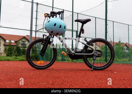 Graues Kinderfahrrad mit blauem Sicherheitshelm und orangen Pedalen, parkt auf rotem Sportplatz neben grünem Zaun. Konzept der Kinderradsicherheit Stockfoto