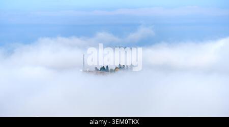Malerischer Blick auf den Hügel mit lebhaftem Herbstlaub, das von sattem Grün bis feurigen Rottönen und goldenen Gelben reicht. Nebelschwaden ziehen unter hellblauem Himmel durch die Landschaft. Stockfoto