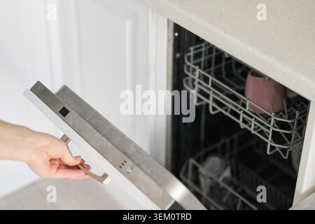Die Geschirrspüler-Tür Wird Von Hand Unter Der Arbeitsplatte Geöffnet Und Zeigt Leeres Untergestell Und Innenraum Aus Edelstahl In Der Weißen Küche. Stockfoto
