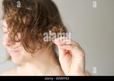 Junge Frau, Die Geschädigtes Haar Untersucht, Kommt Am Ende Aus Nächster Nähe, Während Sie Gespaltene Stränge In Minimalistischer Neutralstellung Für Den Innenbereich Mit Nassem, Lockigem Braun Und Über Den Schultern Liegendem Handtuch Hält. Stockfoto