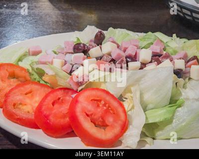 Ein Blick aus der Nähe auf einen frischen Salat, der auf einem weißen Teller serviert wird, mit knusprigem Salat, geschnittenen Tomaten, Schinken- und Käsewürfeln und schwarzen Oliven. Stockfoto