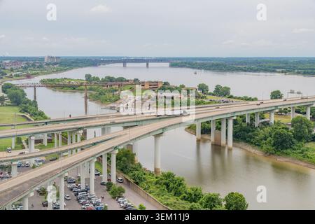 Eine lange Stahlfachwerkbrücke überspannt einen breiten Fluss unter einem bewölkten Himmel, mit einem roten, modernen Gebäude und einer Bootsrampe auf dem grasbewachsenen Ufer. Stockfoto