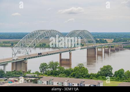 Eine lange Stahlfachwerkbrücke überspannt einen breiten Fluss unter einem bewölkten Himmel, mit einem roten, modernen Gebäude und einer Bootsrampe auf dem grasbewachsenen Ufer. Stockfoto