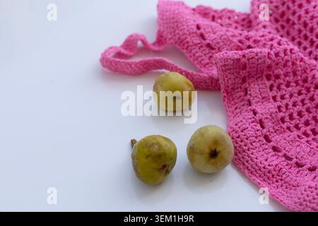 Detailgetreue Aufnahme von drei grünen Feigen, die neben einem hellrosa gestrickten Netzbeutel auf einer glänzend weißen Oberfläche mit Kopierraum angeordnet sind. Stockfoto