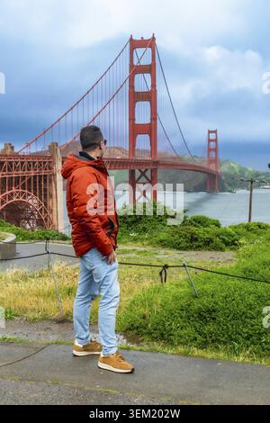 Ein Mann mit orangefarbener Jacke und blauer Jeans steht auf einem Wanderweg und blickt auf die berühmte goldene Gate Bridge, die die Bucht mit Hügeln überspannt Stockfoto
