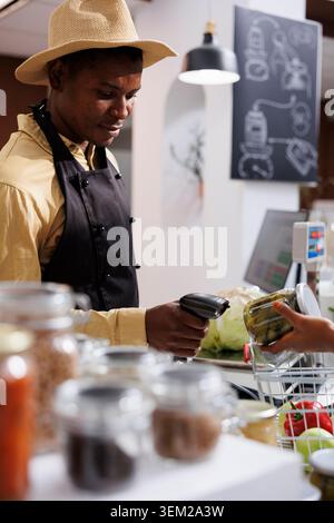 Afroamerikanische männliche Ladenbesitzer in einem umweltfreundlichen Supermarkt fördert nachhaltiges Einkaufen und bietet frische, organische, plastikfreie Produkte an. Detaillierte Aufnahme des Kassierers, der Gegenstände am Tresen scannt. Stockfoto