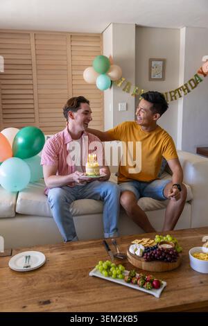 Verschiedene männliche Freunde sitzen auf einem hellen Sofa und haben Geburtstag mit Kuchen, zündeten Kerzen, Ballons, Banner Stockfoto