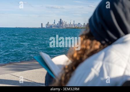 Eine besinnliche Aufnahme einer Person mit langen lockigen Haaren, die einen gemütlichen Hoodie und eine Jacke trägt und auf einer schwarzen Metallbank am Wasser sitzt. Sie Stockfoto