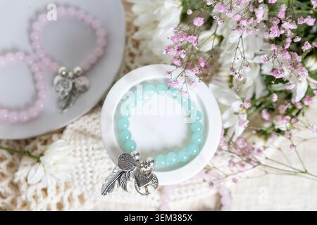 Aus nächster Nähe sehen Sie ein mintgrünes Perlenarmband mit silbernen Reizen in einer weißen Marmorschale, eingerahmt von Blumen. Stockfoto