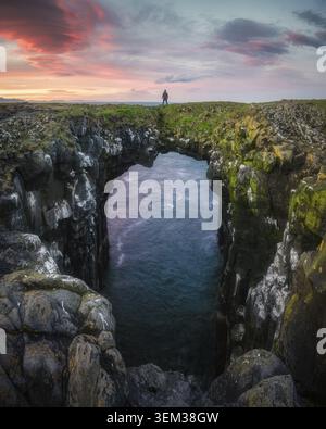 Blick auf eine Person, die auf dem Steinbogen der Gatklettur über dem Meer unter einem dramatischen Sonnenuntergangshimmel mit rosa Wolken in Arnarstapi, Snaefellsbaer, Island steht Stockfoto