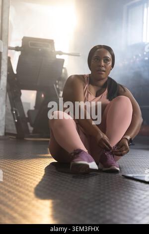 Afroamerikanerin sitzt auf dem Fitnessraum und bindet lila Schnürsenkel mit rosa Set und Smartwatch Stockfoto