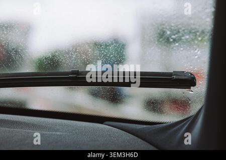 Eine Nahaufnahme aus dem Armaturenbrett eines Fahrzeugs mit Blick auf ein schwarzes Wischerblatt, das auf einer regenbedeckten Windschutzscheibe mit einem verschwommenen Straßenrand geparkt ist Stockfoto