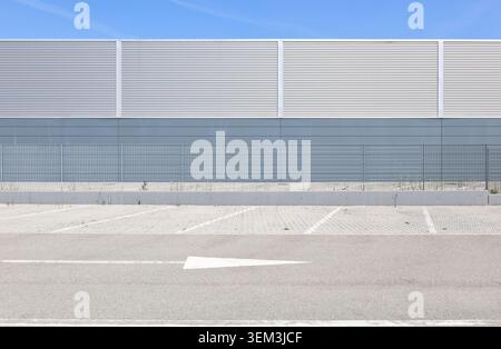Metallgitterzaun mit einem modernen Industriebau dahinter. Straße, Parkplatz und Straßenmarkierung davor. Hintergrund für Kopierbereich. Stockfoto