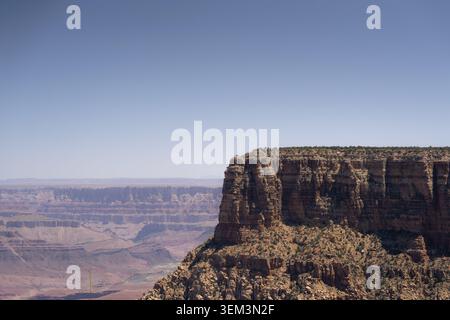 Felsiger Klippenrand mit Blick auf eine riesige Wüstenschlucht unter einem klaren blauen Himmel, mit mehrschichtigen Steinmauern und weit entfernten Bergrücken in sanftem Sonnenlicht. Stockfoto
