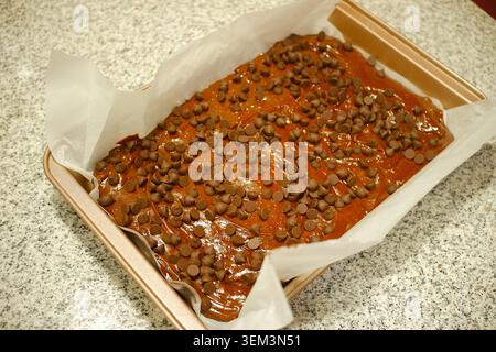 Ein roher Brownie-Teig, großzügig mit Schokoladenchips belegt, bereit zum Backen in einer mit Pergamentpapier ausgelegten, rechteckigen Pfanne. Stockfoto