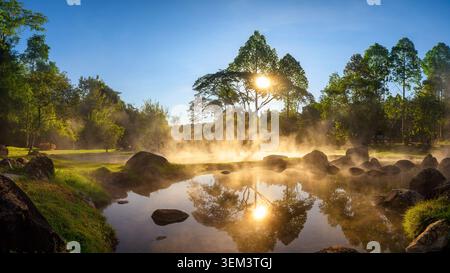 Heitere heiße Quelle während des Sonnenaufgangs, mit Nebel über reflektierendem Wasser, umgeben von Felsen, Bäumen und üppigem Grün. Das goldene Sonnenlicht erzeugt Stockfoto
