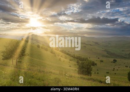 Malerischer Sonnenaufgang mit Sonnenstrahlen, die durch Wolken über sanfte grüne Hügel und verstreute Bäume in der Nähe des Berges Tschyortow Palets, Altai Republik, Russi Stockfoto