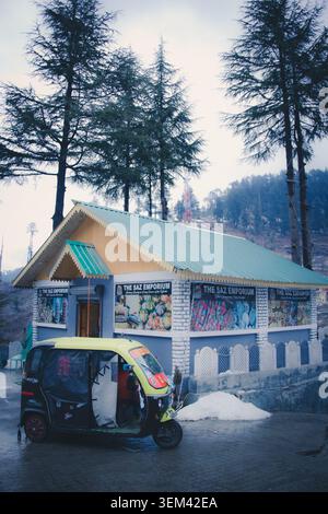Eine dreirädrige Auto-Rikscha parkt vor einem rustikalen Gebäude, umgeben von hohen Kiefern in Kaschmir, Indien. Stockfoto