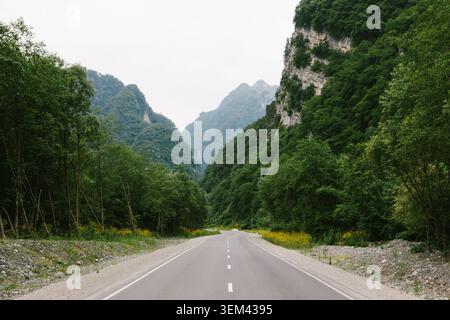 Leere Asphaltstraße, die in die Karmadon-Schlucht führt, Nordossetien–Alania, Russland, umgeben von grünen bewaldeten Bergen und steilen Felsklippen auf einem cl Stockfoto