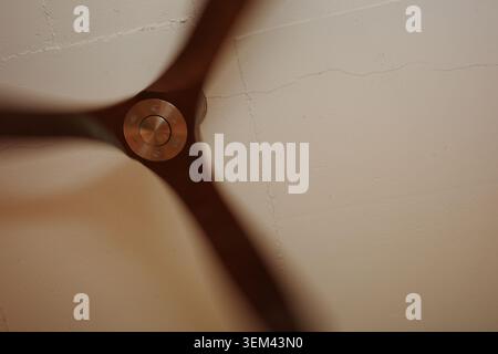 Nahaufnahme eines sich drehenden Deckenventilators aus Holz mit einer metallischen Nabe in Bewegung, die unscharf gegen eine rissige Putzdecke ist, warme Innenbeleuchtung. Stockfoto