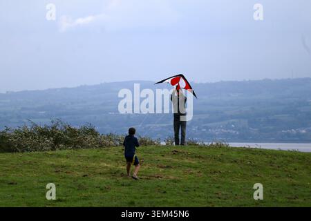 Ein Vater und sein Sohn fliegen einen rot-schwarzen Stunt-Drachen auf einem grasbewachsenen Hügel mit Blick auf die Dee-Mündung und die Küste von Nordwales in Thurstaston. Wirral, UK. Stockfoto