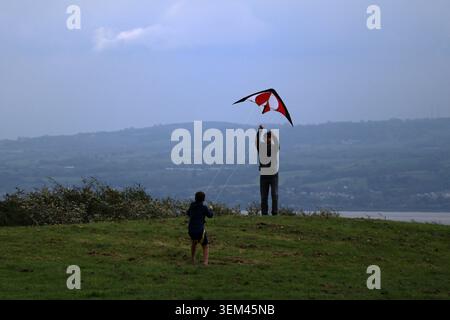Ein Vater und sein Sohn fliegen einen rot-schwarzen Stunt-Drachen auf einem grasbewachsenen Hügel mit Blick auf die Dee-Mündung und die Küste von Nordwales in Thurstaston. Wirral, UK. Stockfoto