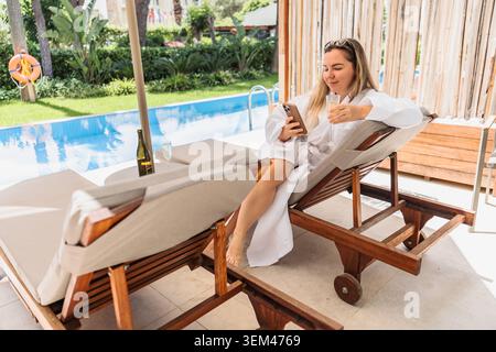 Frau im Bademantel, die sich am Pool mit ihrem Telefon entspannt Stockfoto